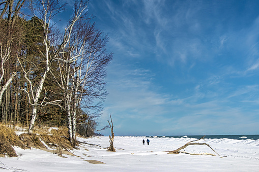 Beneath a blue sky with white clouds on a winter's day, a couple walks off into the distance on the frozen shoreline of Lake Michigan at Harrington Beach near Lake Church, WI.