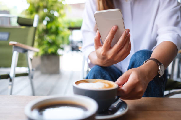 istock Closeup image of a young woman holding and using mobile phone while drinking coffee with friend in cafe 1392251790