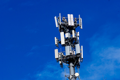 A communications tower with multiple cellular antennae and related tele-communications equipment stands out against a blue sky.