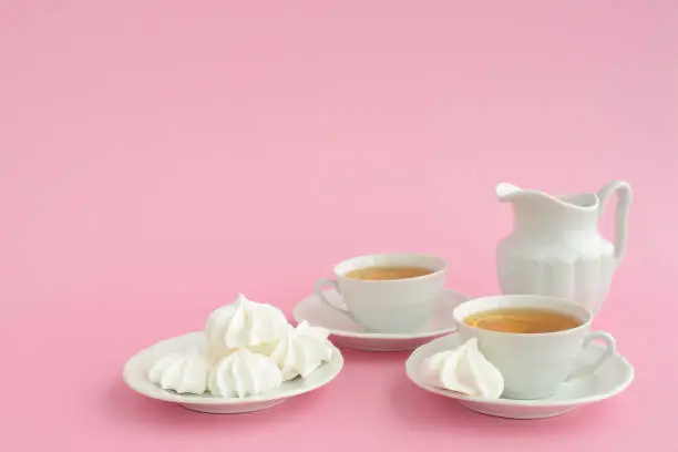 Photo of Tea and marshmallows. Black tea with lemon in white porcelain cups, porcelain milk jug and sweets on a pink background