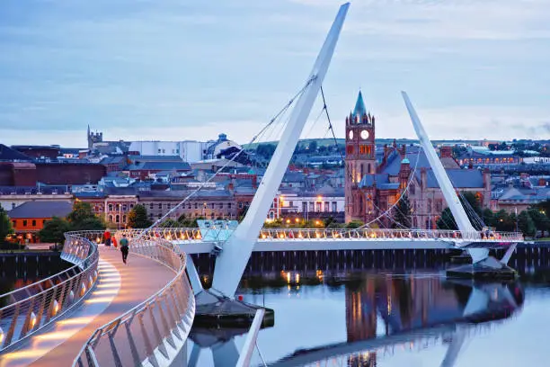 Photo of Derry, Ireland. Illuminated Peace bridge in Derry Londonderry, City of Culture, in Northern Ireland with city center at the background. Night cloudy sky with reflection in the river at the dusk