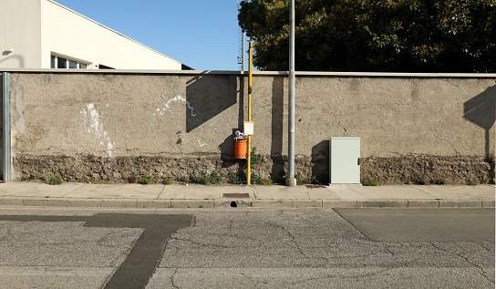 Yellow pole with small blank sign and full waste bin. Grunge concrete wall on behind, sidewalk and weathered asphalt road in front. Background for copy space.