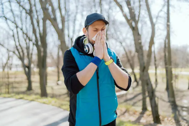 Photo of A man with a pollen allergy stands outside in a park. An athlete stops running practice due to a runny nose, blows his nose in a handkerchief. Illness interferes with exercise