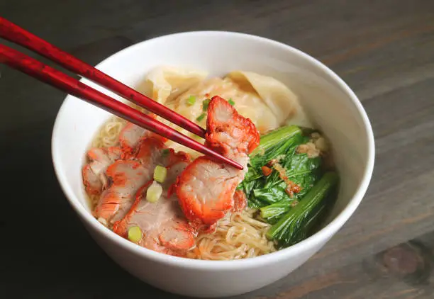 Photo of Bowl of Mouthwatering Egg Noodle and Wonton Dumpling Soup with Roasted Pork on Wooden Table