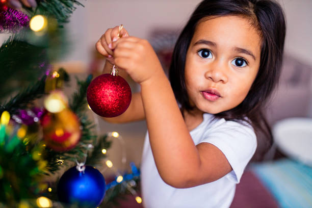 gemischtrassiges kleines süßes mädchen, das am weihnachtsmorgen im schlafzimmer einen weihnachtsbaum schmückt und einen wunsch wünscht - 1 2 months stock-fotos und bilder