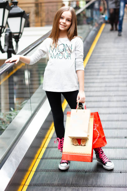 criança elegante em boina francesa e casaco moderno. menina com sacolas de compras. tempo da primavera. tendência de outono. dia de compras. vida feliz moderna. garotinha. look de moda infantil. copiar espaço. vida moderna. - escalator child shopping mall little girls - fotografias e filmes do acervo