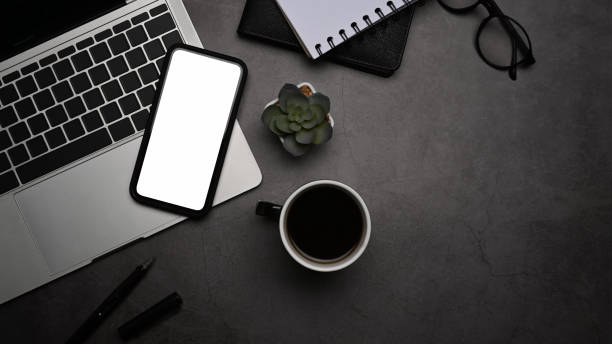 istock Flat lay smart phone, laptop computer, coffee cup, notepad and glasses on dark background. 1392278201