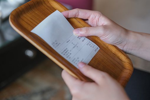 Close-up shot of an unrecognizable customer checking the restaurant bill before paying for her lunch. She's holding the bill on top of a small wooden tray.