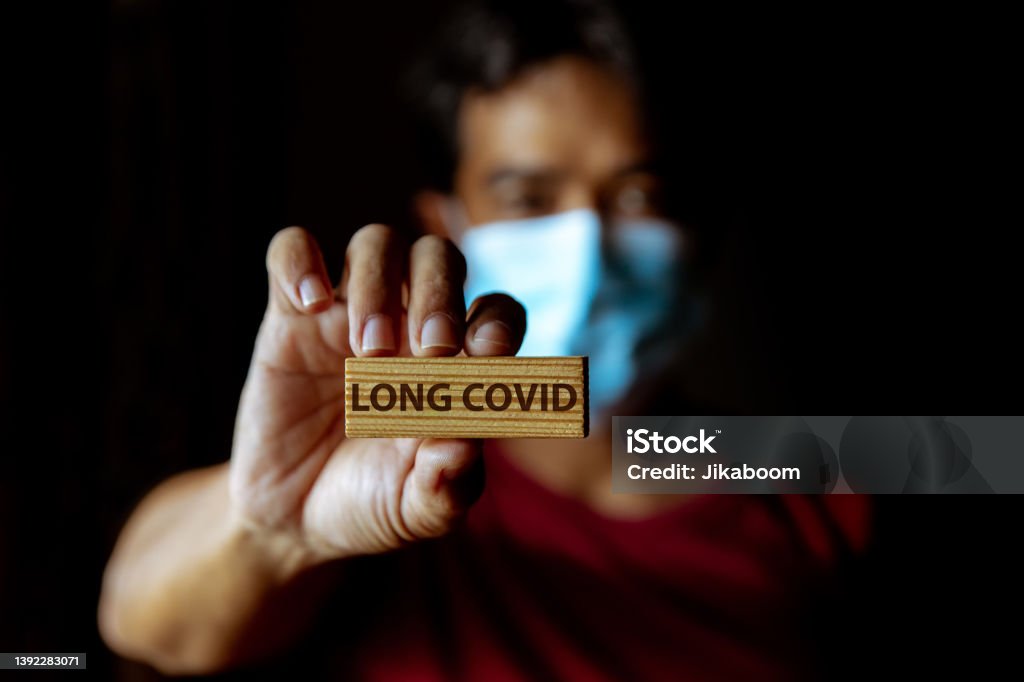 Masked Asian man show wooden sign with wording "Long Covid" Masked Asian man show wooden sign with wording "Long Covid" in dark room. Long COVID Stock Photo