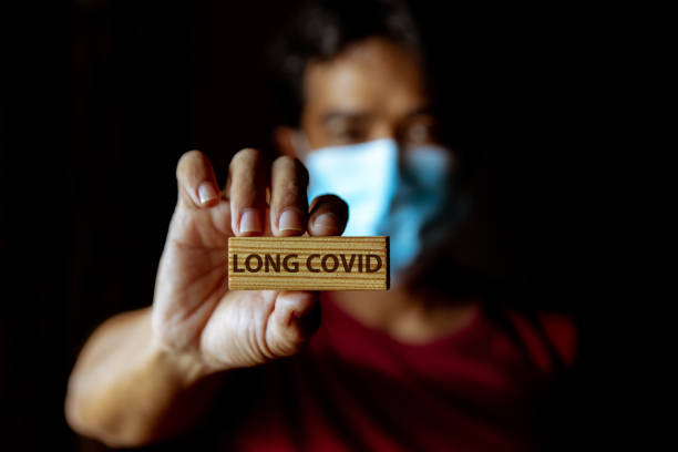 Masked Asian man show wooden sign with wording "Long Covid" Masked Asian man show wooden sign with wording "Long Covid" in dark room. long covid stock pictures, royalty-free photos & images