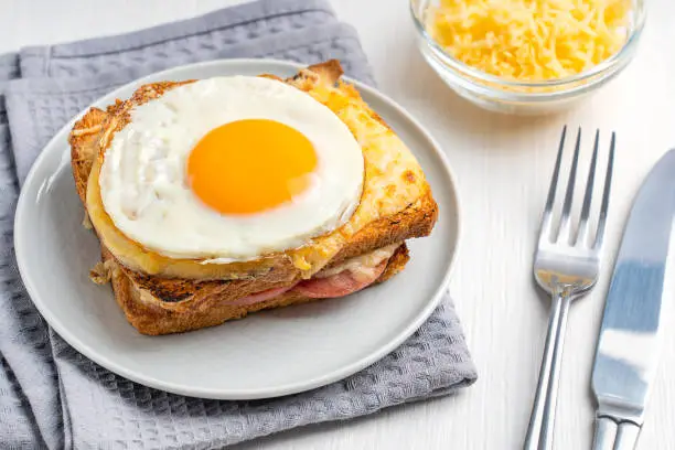 French Croque Madame local cuisine a hot sandwich made with ham and cheese inside toasted bread with topping of fried egg served on plate with tableware and towel on white wooden table for breakfast