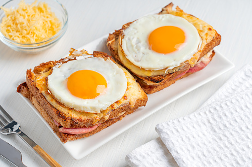Two Croque Madame French local hot sandwiches made with ham and cheese inside toasted bread topped with fried egg with yolk served on plate with tableware and towel on white wooden table for breakfast