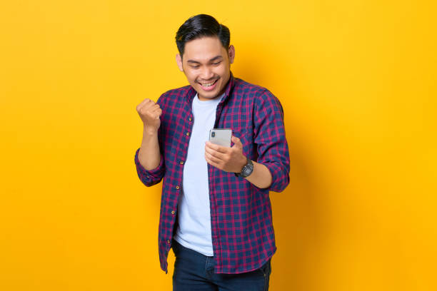 istock Excited young Asian man in plaid shirt using smartphone and doing winner gesture isolated on yellow background 1392295895
