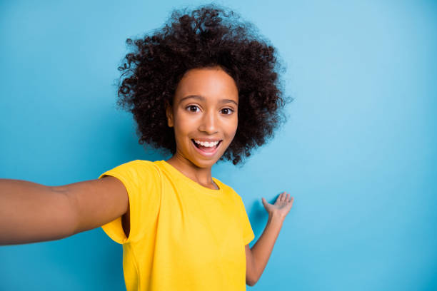 Photo of cheerful little afro american girl wear yellow t-shirt invite hand make selfie isolated on blue color background stock photo