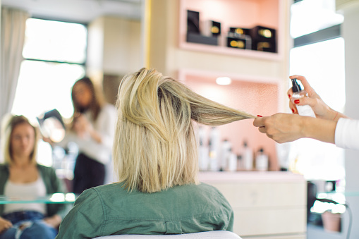 Woman patiently sitting in hair salon while hairdresser finishes hair styling by applying hair spray.