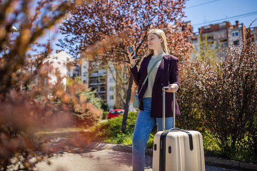 Young Caucasian woman, using mobile phone, while travel with suitcase