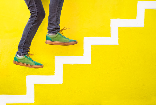 Side view cropped shot of man feet wearing sneaker pretending to walk up on painted steps on yellow wall