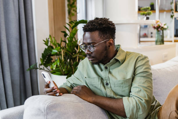 istock Young African-American man using mobile phone while sitting on sofa at home 1392301045
