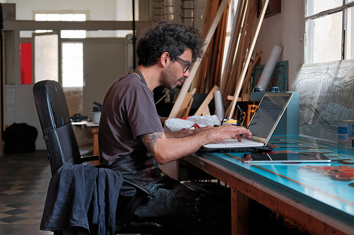 A man sits at a desk and uses a laptop in an art and craft studio.