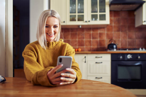 istock Smiling mature woman using mobile phone at home sitting in the kitchen 1392301418