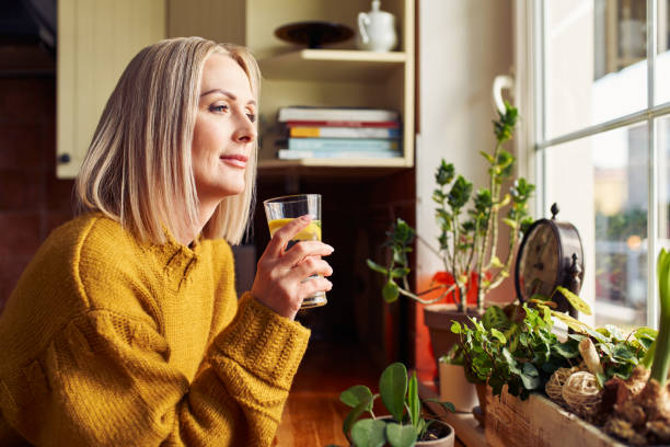 donna matura che beve un bicchiere d'acqua in cucina guardando attraverso la finestra - foods and drinks simplicity purity clothing foto e immagini stock