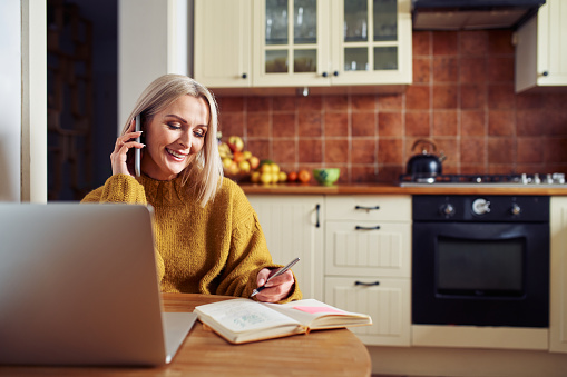 Smiling mature woman sitting in the kitchen at home talking on the phone using laptop writing in notebook
