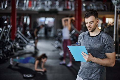 Young fitness instructor writing sports training plans in a gym. There are people in the background.