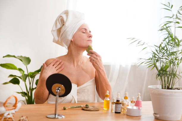 smiling woman 35 year plus in the towel using gua sha stone for facial massage. Cosmetic natural trend. Morning hygiene routine and skincare treatment stock photo