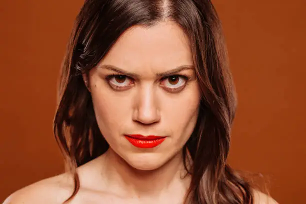 Face portrait of young angry hateful woman looking at camera at studio over brown background. Upset, frustrated, anger, rage female facial expression.