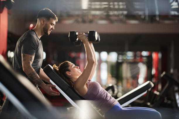 Young man helping his girlfriend during her sports training in a health club. Fitness instructor assisting athletic woman in exercising with dumbbells at gym. gym stock pictures, royalty-free photos & images