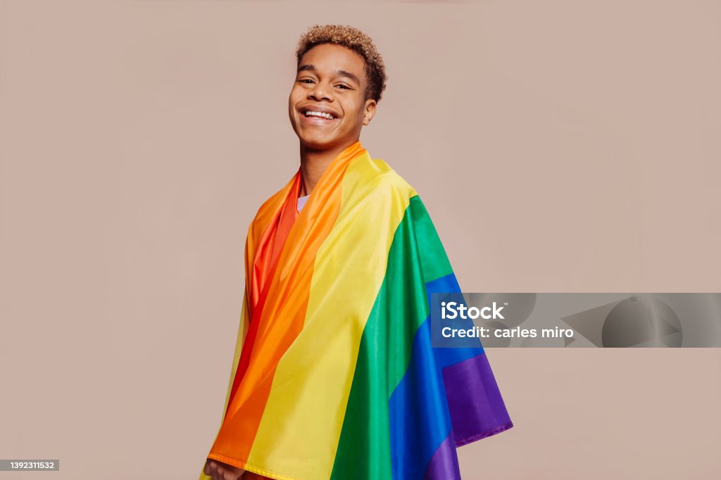 Cheerful latin american young man with a rainbow flag Portrait of happy smiling afro latin american young man with a gay pride rainbow flag at studio over beige background. Homosexual lgbtiq concept, rainbow flag, celebrating parade. Copy space. LGBTQIA Rights Stock Photo