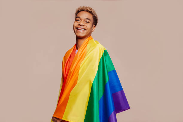 Cheerful latin american young man with a rainbow flag Portrait of happy smiling afro latin american young man with a gay pride rainbow flag at studio over beige background. Homosexual lgbtiq concept, rainbow flag, celebrating parade. Copy space. gay pride people stock pictures, royalty-free photos & images