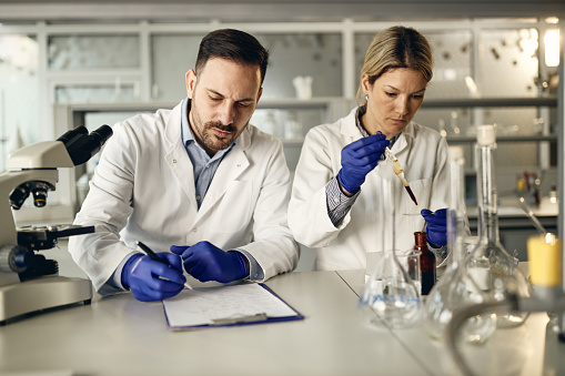 Two chemists working on medical research in laboratory. Focus is on man taking notes.