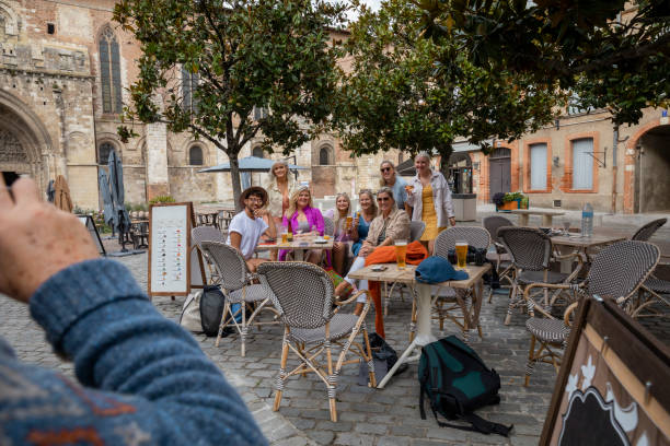 An over-the-shoulder view of a medium group of friends enjoying a refreshment break whilst on a trip away to Toulouse in the South of France. They are having their picture taken by one of the men in thr group.