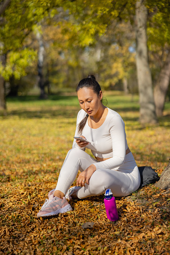 Asian woman in casual sports clothes sitting on dry leaves in the forest, looking at smart phone and relaxing after workout outdoors