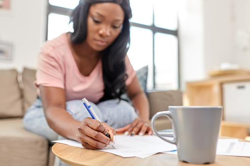 accounting, taxes and finances concept - young african american woman with papers and calculator at home