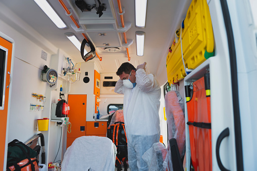 First aider healthcare emergency young doctor man or frontline worker dressing white protective medical disposable hooded coveralls suit with glove, face shield and N95 face mask gear Preparing for emergency response to the patient from the medical service ambulance car during the covid19 pandemic after coronavirus quarantine and lockdown.