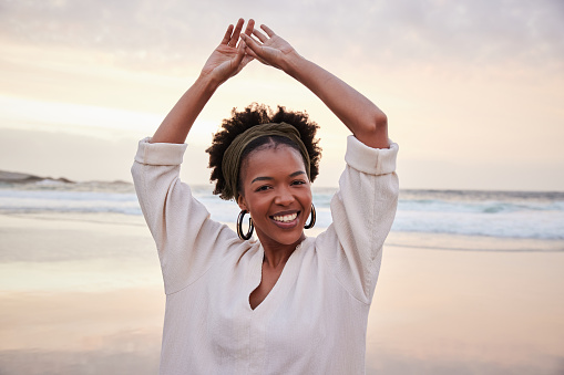 Portrait of a smiling young African woman standing with her arms in the air on a sandy beach at sunset