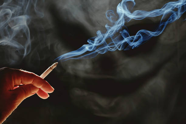 istock Man's hand holding cigarette with white smoke look like devil face on dark background 1392325302