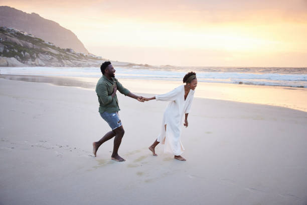 pareja joven riendo mientras corren juntos por una playa de arena al atardecer - luna de miel fotografías e imágenes de stock