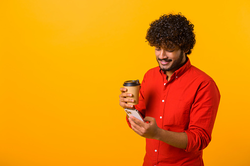 Confident middle eastern man wearing smart casual shirt texting on the smartphone and holding a paper cup of take-away coffee. Male office employee isolated on orange