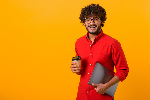 Waist up photo of handsome cheerful smiling Indian man extends hand, holds takeaway coffee cup, drinks coffee or tea, smiling to the camera isolated over orange background