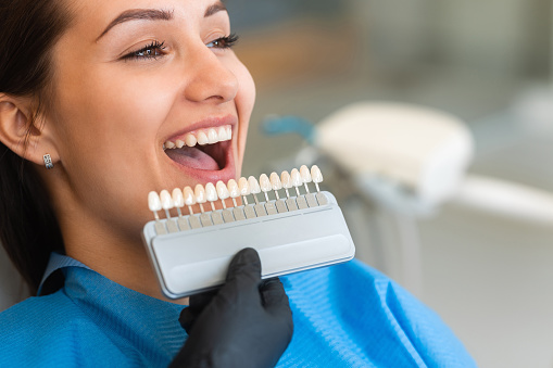 A beautiful young patient uses a dental tooth shade guide for whitening teeth service to ensure a perfect choice of color. Bleaching, prosthetics, orthopedic dentistry concept.