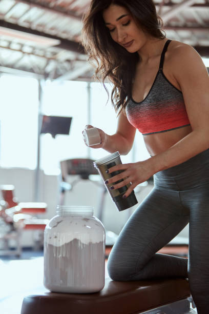 A woman is making a protein shake after a workout in the gym. She uses a measuring cup to mix the protein powder in her cup. stock photo