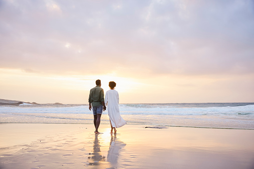 Rear view of a young African couple holding hands and walking along a sandy beach together at sunset