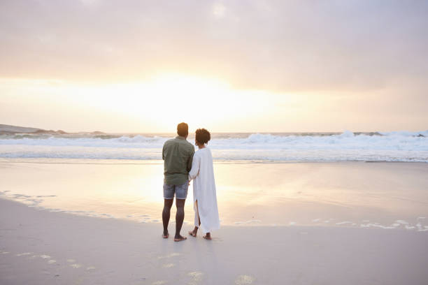 istock Young couple walking holding hands on a beach and watching the sunset 1392328170