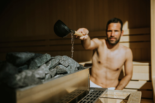 Handsome young man pouring water onto hot stone in the sauna