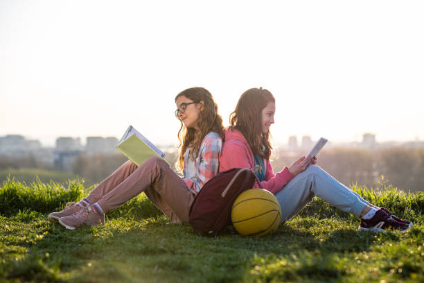 自然の中に座っている二人の少女 一人はタブレットを見つめ、もう一人は本を読んでいる - basketball teenager nature outdoors ストックフォトと画像