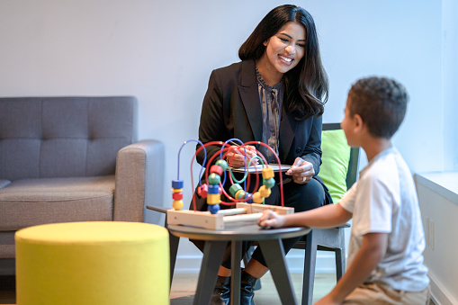 A female therapist sits in a chair beside her young patient as they play together with a bead maze. The therapist is dressed professionally in a suit and has a tablet out on her lap to take notes about the session. The young causally dressed boy is kneeling on the floor n front of the toy as they talk together.