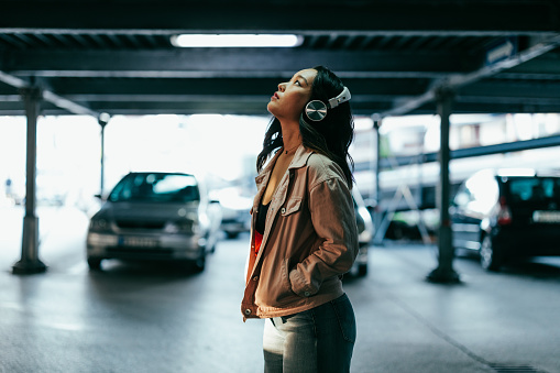 Portrait of asian young pretty and attractive woman looking up while standing at a car parking lot with earphones on